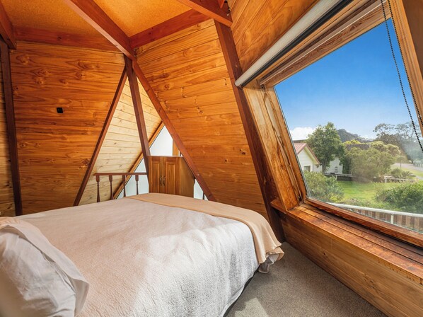 A bedroom with a sloped ceiling, wooden walls, a bed with a white comforter, and a window overlooking a landscape.