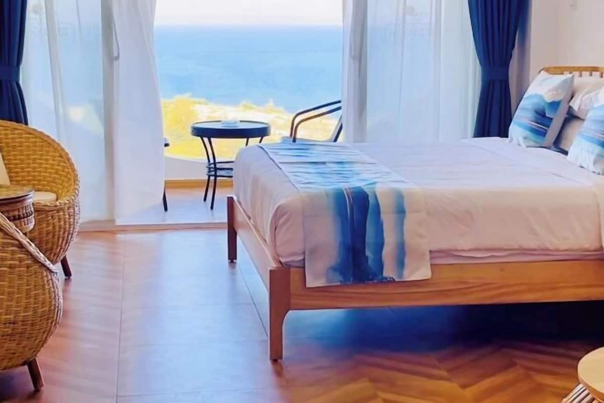 A bedroom with a bed, a chair, a small table, and a view of the ocean.