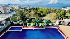 Une piscine divisée en deux zones, entourée de chaises longues et de parasols, et offrant une vue sur une zone côtière avec des bâtiments et des palmiers.