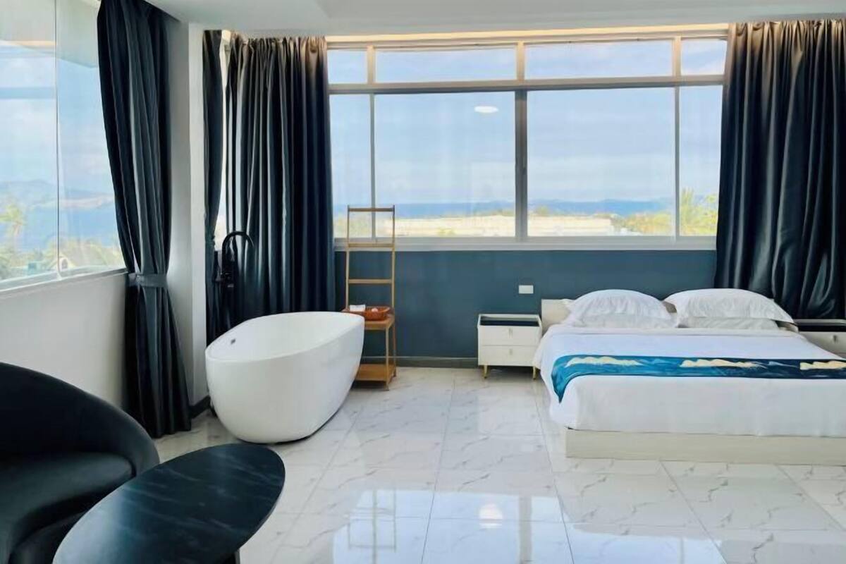 A modern bedroom with a large bed, a white bathtub, a chair, and a view of the outdoors.