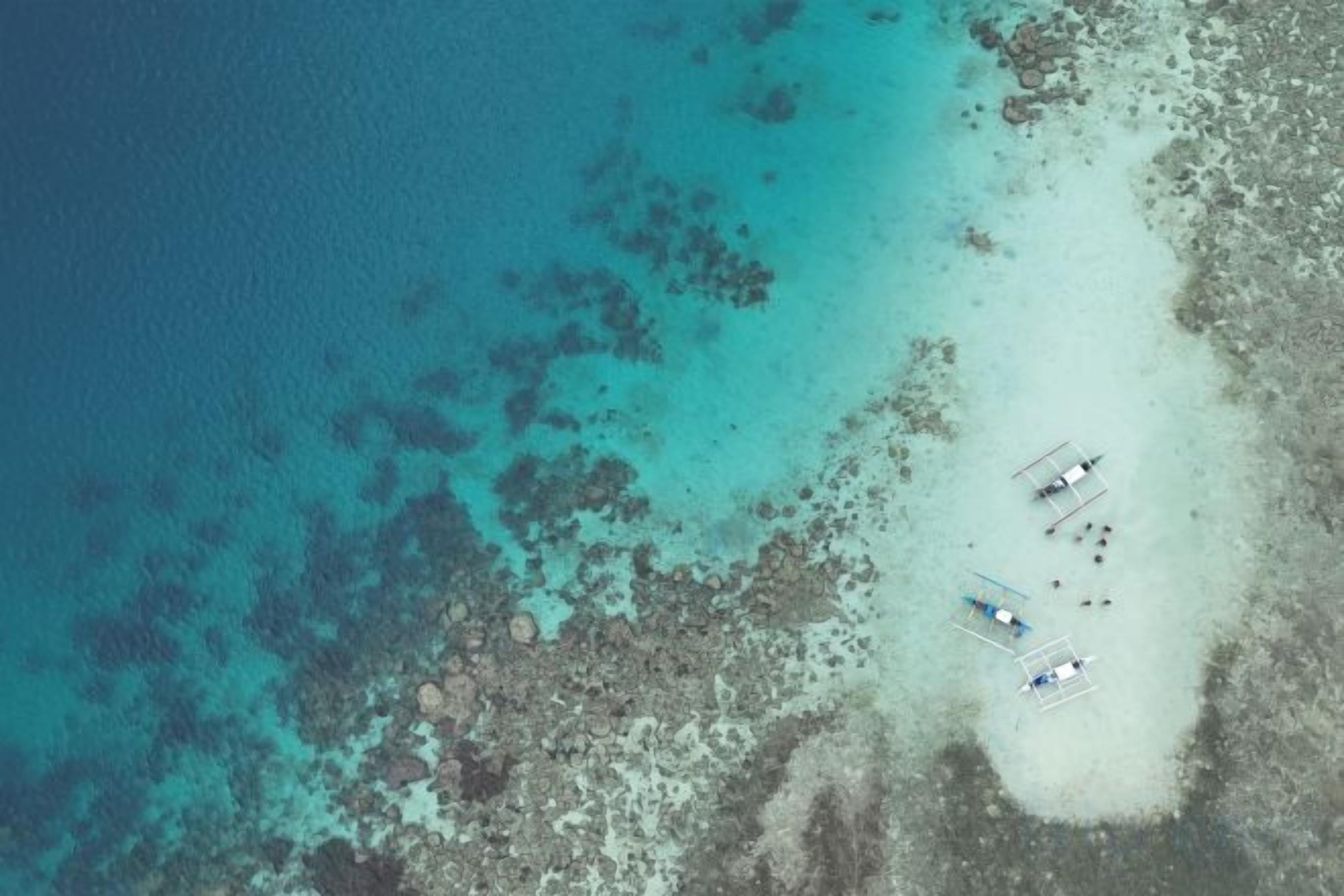 An aerial view of a beach with clear turquoise waters and a sandy shore. There are several boats anchored near the shore.