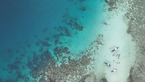 Vue aérienne d’une plage aux eaux turquoise et limpides, bordée d’une côte rocheuse et où plusieurs petits bateaux sont amarrés près du rivage.