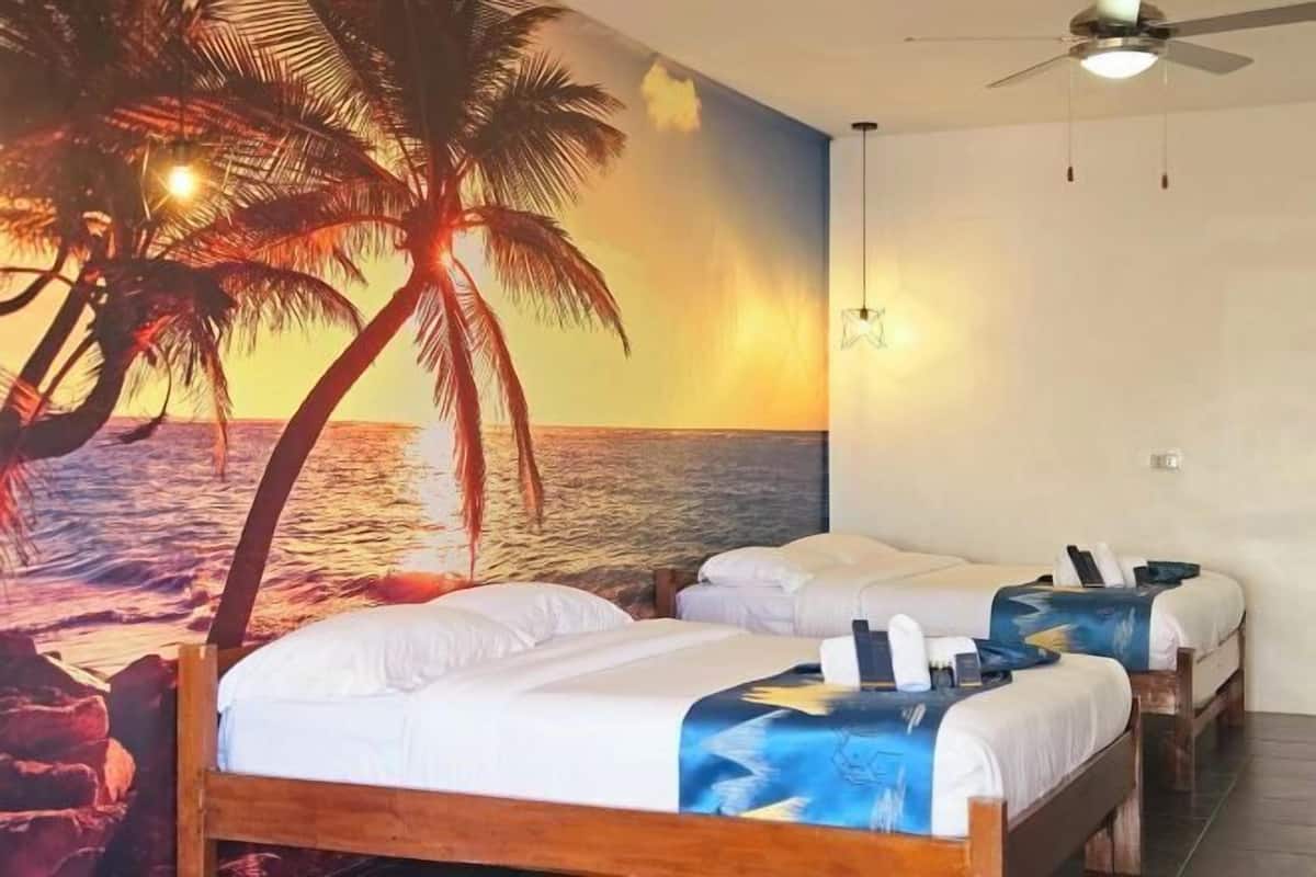 A room with two beds, a ceiling fan, and a mural of a sunset over the ocean.