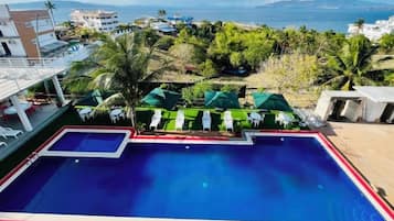 A swimming pool with a view of the sea and mountains, surrounded by greenery and lounge chairs.