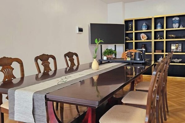 A dining area with a long table, chairs, a plant, a flat-screen TV, and a shelf with decorative items.