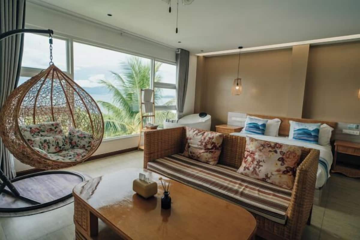 A room with a hanging chair, a sofa with floral cushions, a wooden coffee table, and a view of palm trees through large windows.