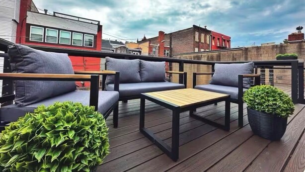 A rooftop patio with a wooden deck, grey sofas, a wooden table, and a potted plant.