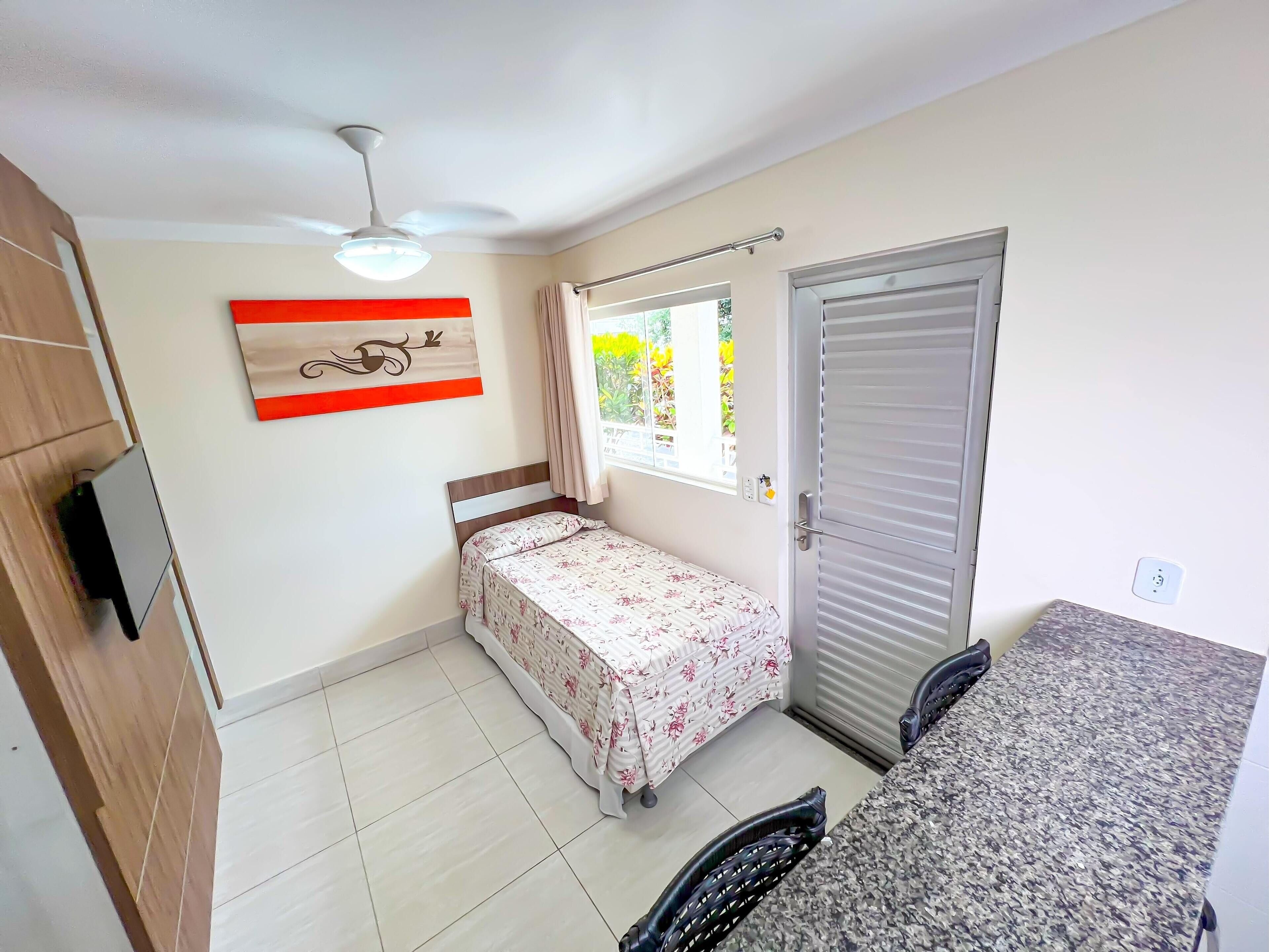 A small bedroom with a bed, a ceiling fan, a window with curtains, a door, and a small kitchenette area.