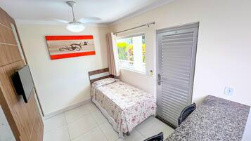 A small bedroom with a bed, a ceiling fan, a window with curtains, a door, and a small kitchenette area.