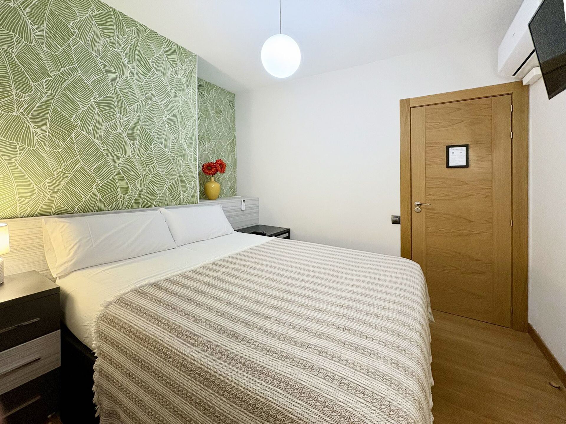 Quarto com cama, mesa de cabeceira, luminária na parede, porta de madeira e papel de parede estampado.