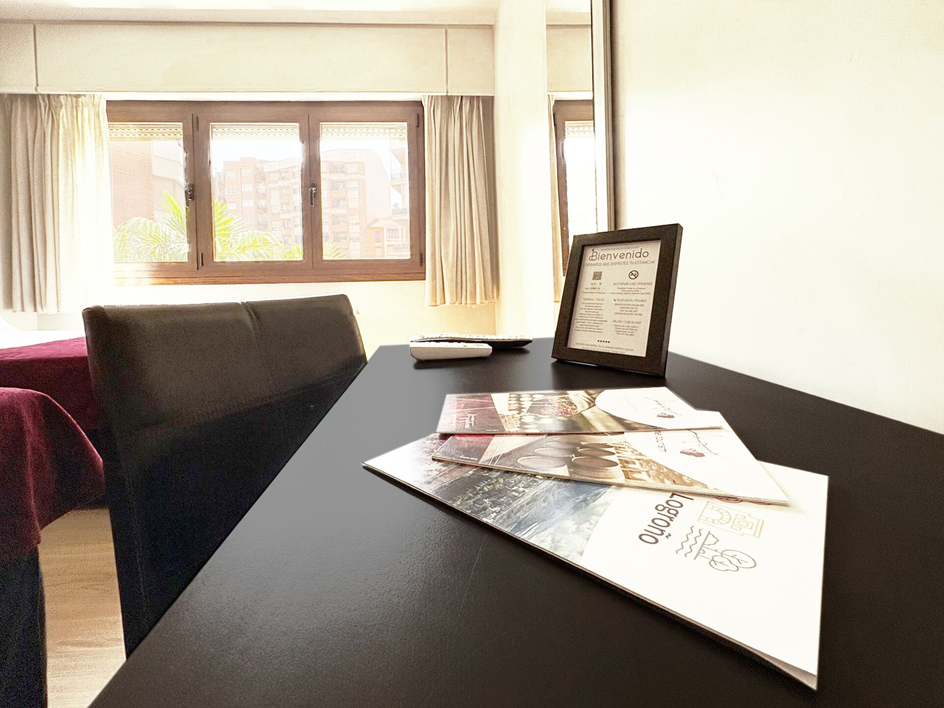 Habitación de hotel con una mesa negra, un cartel de bienvenida enmarcado y un ventanal con cortinas.