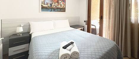 A hotel room with a bed, a nightstand, a lamp, a painting on the wall, and a view of the outside through a door.