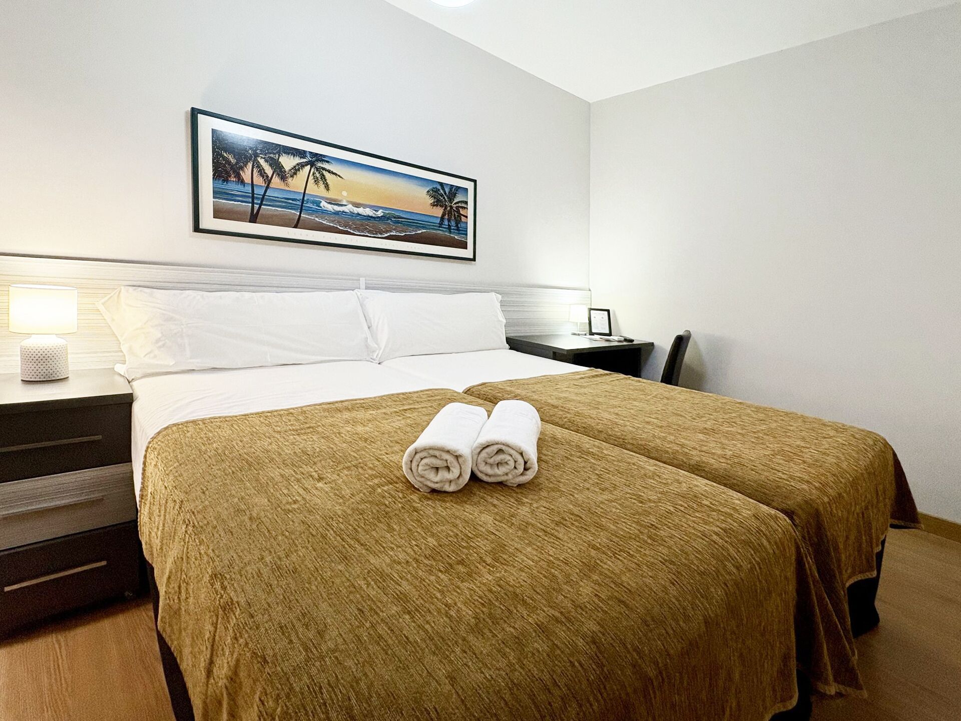 A hotel room with a bed, two pillows, a nightstand with a lamp, a framed picture on the wall, and a pair of rolled towels on the bed.