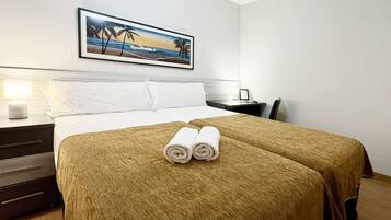 A hotel room with a bed, two pillows, a nightstand with a lamp, a framed picture on the wall, and a pair of rolled towels on the bed.