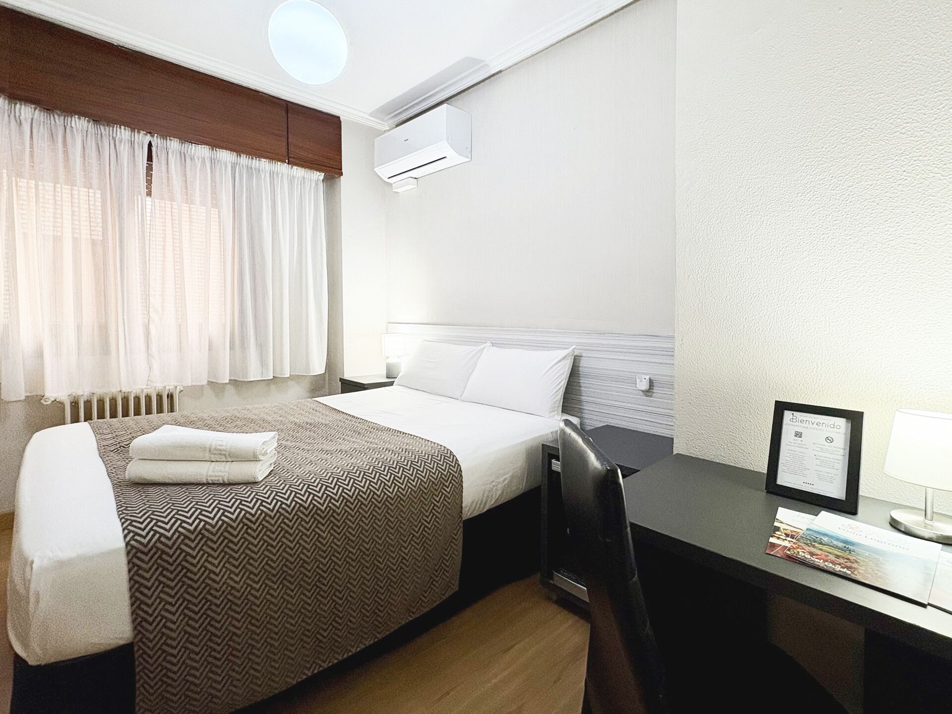 Ein Hotelzimmer mit Bett, Schreibtisch, Stuhl, Klimaanlage und einem fenster mit Vorhängen.