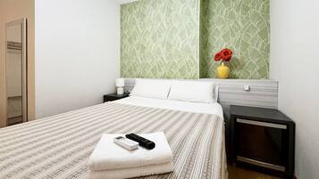 Une chambre d’hôtel avec un lit, une table de chevet, une lampe, une télécommande et un vase avec des fleurs.