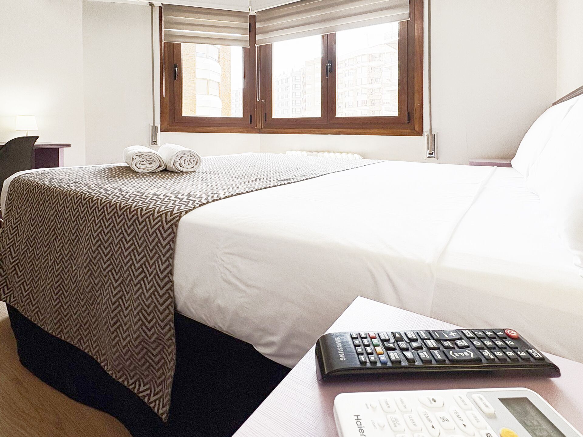 A hotel room with a bed, a desk with a Samsung remote control, and a white alarm clock.