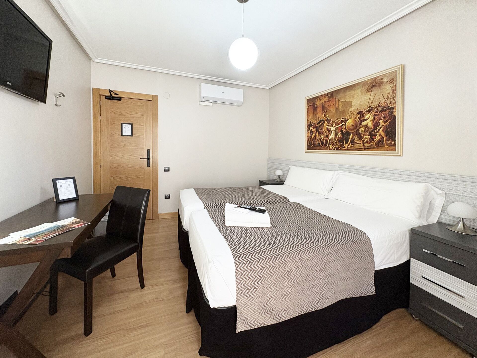 A hotel room with a bed, a desk, a chair, a nightstand, and a painting on the wall.