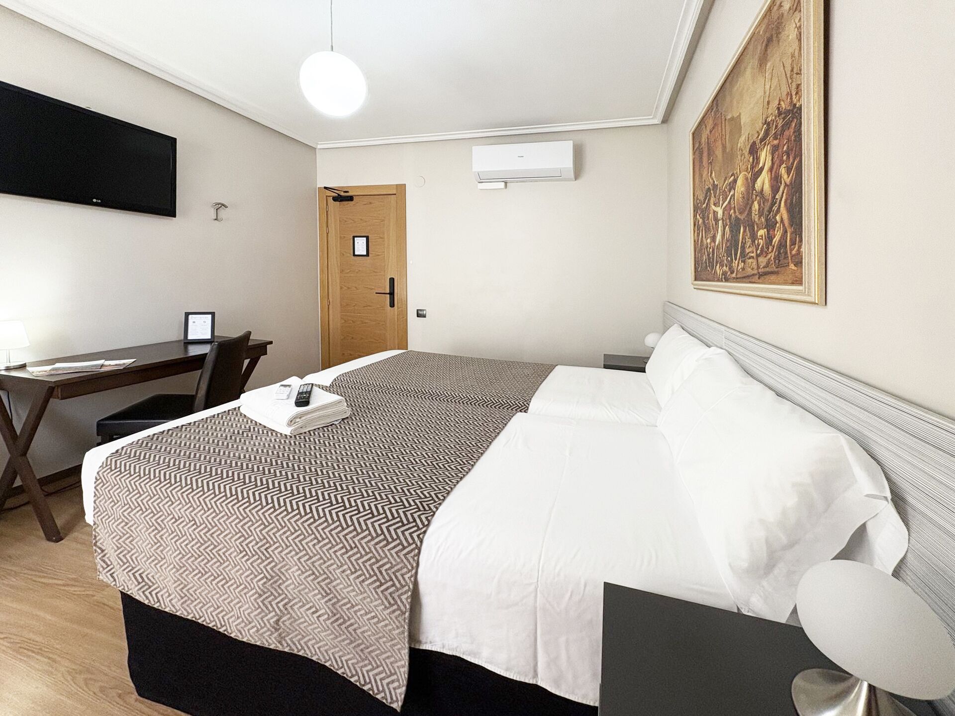 Habitación de hotel con cama, escritorio, silla, televisor y un cuadro en la pared.