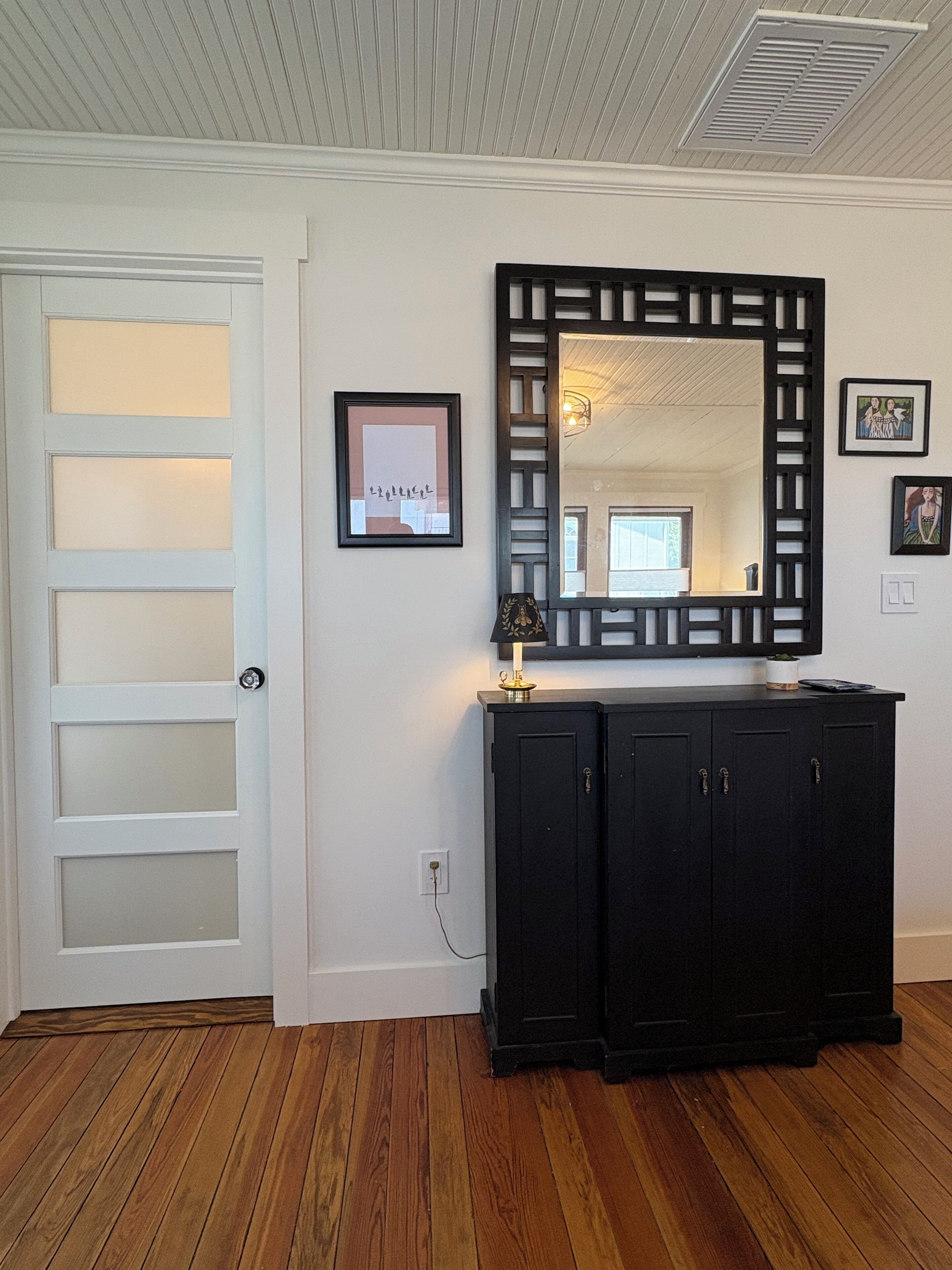 A room with a white door with frosted glass panels, a black cabinet with a mirror above it, and a framed picture on the wall.