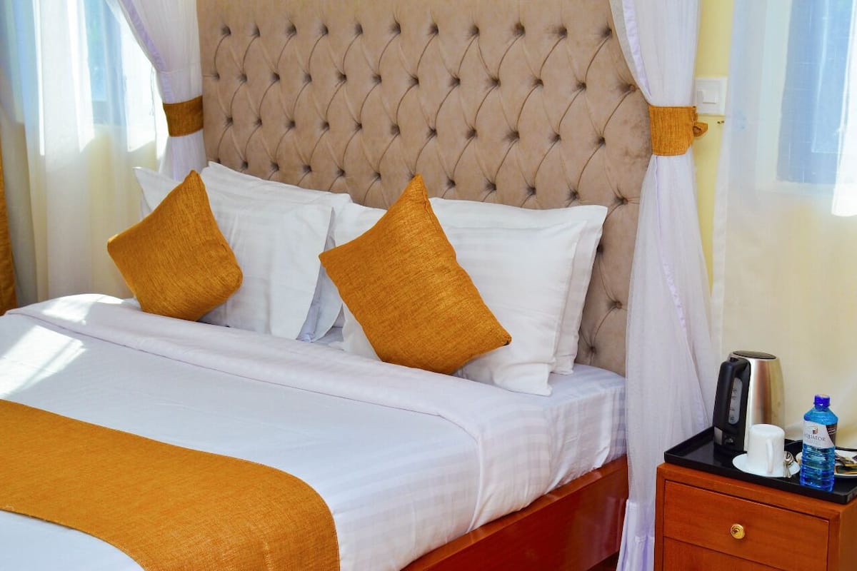 A neatly made bed with white and mustard pillows, a tufted headboard, and a bedside table with a water bottle and a coffee maker.
