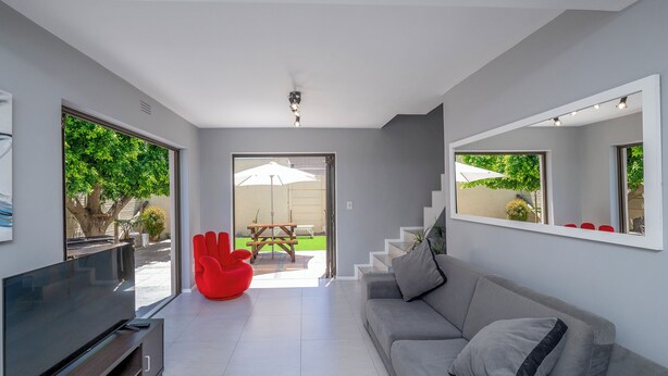 A modern living room with a flat-screen TV, grey sofa, red chair, and a staircase leading to an outdoor area with a picnic table and umbrella. - Xanadu (Cape Town)