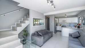 A modern living room with a grey sofa, a glass coffee table, and a staircase leading to another level. - Xanadu (Cape Town)