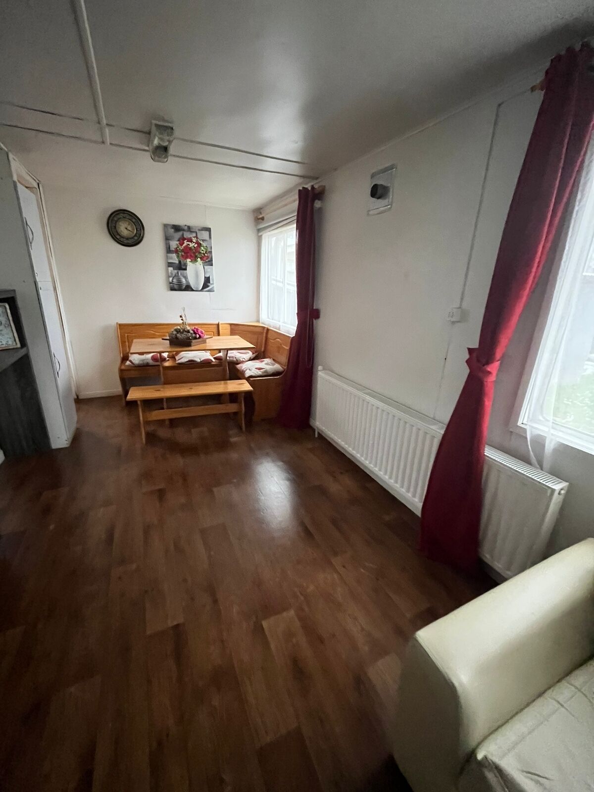 A room with a wooden floor, a bed with a headboard, a table with chairs, a radiator, and a window with curtains.
