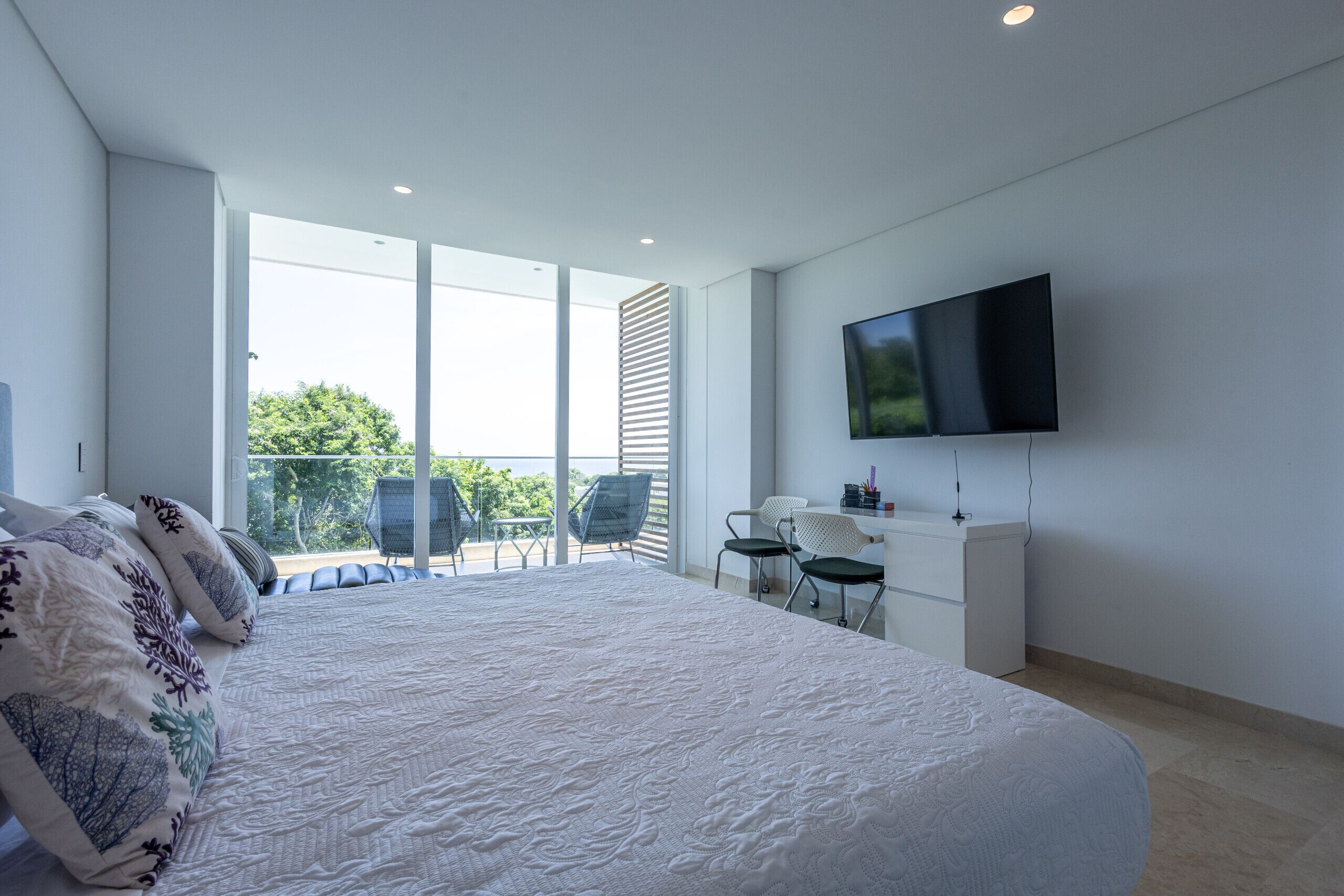 A modern bedroom with a large bed, a TV mounted on the wall, and a balcony with chairs.