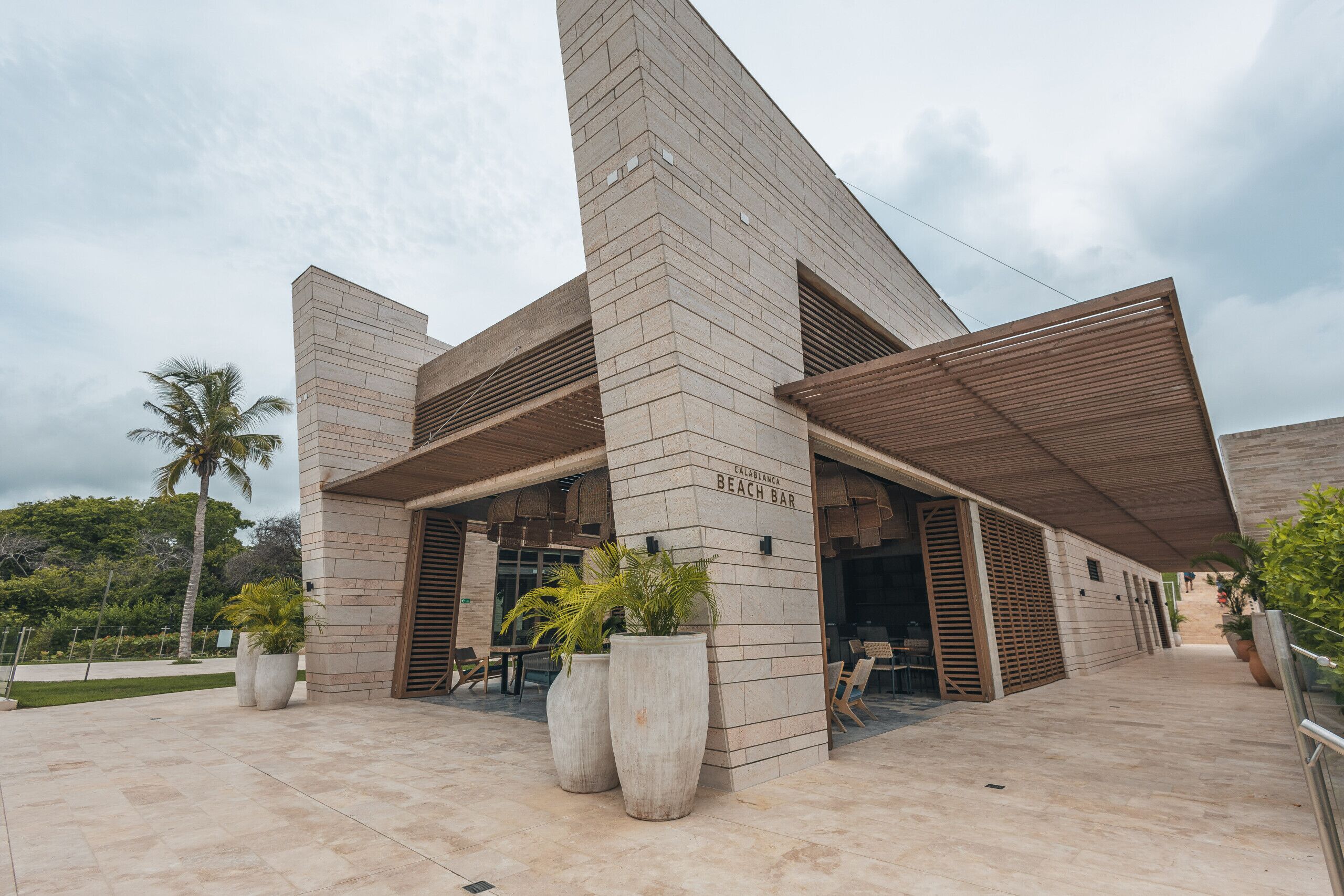 A modern building with a prominent entrance, large potted plants, and a sign indicating a beach bar.