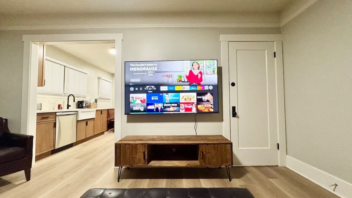 A living room with a flat-screen TV mounted on the wall, a wooden coffee table, and a black leather sofa.