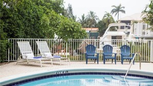 A swimming pool with lounge chairs and a small table, surrounded by a white fence and greenery.