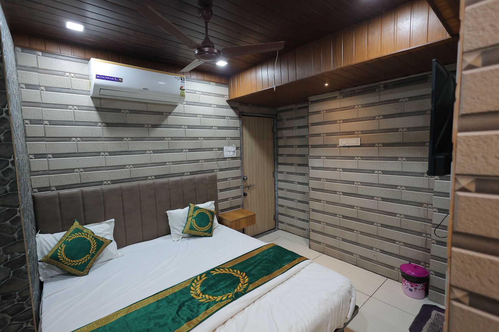 Une chambre avec un mur décoré d’un motif en briques, un lit avec une literie blanche et des coussins verts et dorés, un ventilateur de plafond et une petite table de chevet.