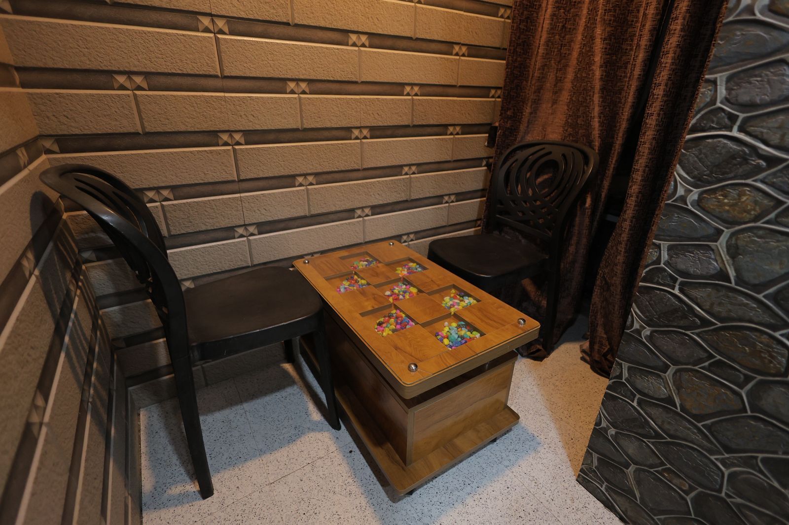 Una pequeña mesa de madera con superficie a cuadros y bolas de colores, ubicada entre dos sillas negras en una habitación con pared de ladrillo y piso de piedra.