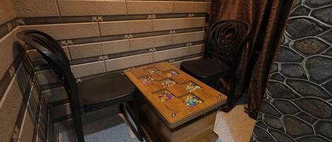Una pequeña mesa de madera con superficie a cuadros y bolas de colores, ubicada entre dos sillas negras en una habitación con pared de ladrillo y piso de piedra.