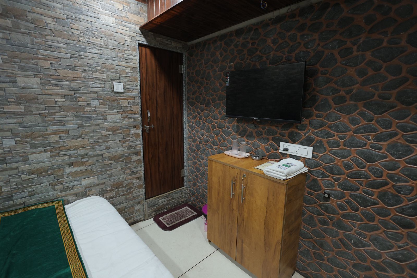 A room with a stone wall, a wooden door, a bed with a green and white bedspread, a wooden cabinet, and a flat-screen TV mounted on the wall.