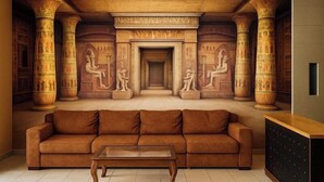 A room with a brown sofa, a glass coffee table, and a mural of Egyptian columns and figures. - The Great Golden Pyramid (Giza)