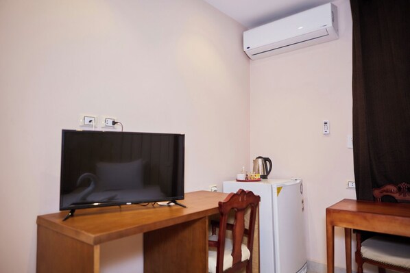 Une petite pièce comprenant un bureau en bois, un téléviseur, un réfrigérateur blanc et un appareil de climatisation.