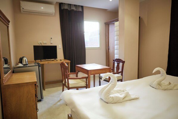 A hotel room with a bed, two wooden chairs, a small desk with a computer and a kettle, and a window with curtains.