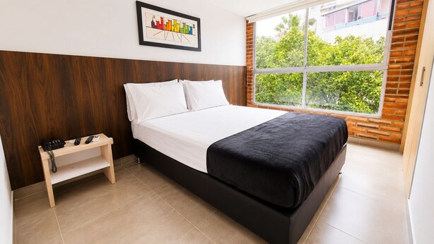 Een hotelkamer met een bed, een nachtkastje met een telefoon, een raam met uitzicht op groen en een stadsgezicht foto aan de muur.