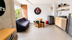 Un espacio habitable compacto con sofá, una pequeña mesa de madera, una zona de cocina con refrigerador y un logotipo montado en la pared.
