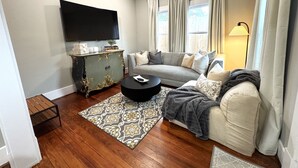 A living room with a grey sofa, a black coffee table, a vintage cabinet, a flat-screen TV, and a window with curtains. - Cozy Houston Retreat • Private Gated | Holly House (Houston)