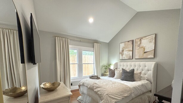 A bedroom with a large bed, a bedside table, a mirror, and a window with curtains. - Cozy Houston Retreat • Private Gated | Holly House (Houston)