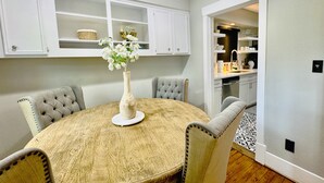 A dining area with a round table, grey upholstered chairs, and a vase with flowers. White cabinetry with shelves is above the table. - Cozy Houston Retreat • Private Gated | Holly House (Houston)