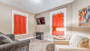 A living room with two windows, a flat-screen TV, a small table with a lamp, and a couch. - Urban Comfort Nook - Walk to Overton Park (Memphis)