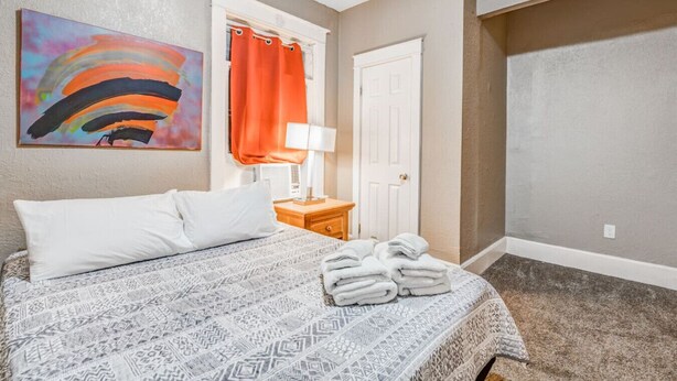 A hotel room with a bed, a painting, a window with orange curtains, a door, and a carpeted floor. - Urban Comfort Nook - Walk to Overton Park (Memphis)
