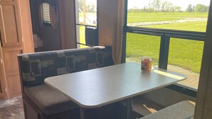 A compact RV interior with a dining area featuring a table and benches, a small kitchen area, and a view of a green field through the window.