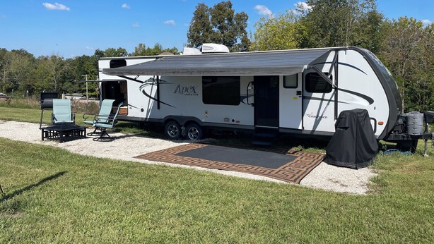 A white RV with "Apex Ultra Lite" branding parked on a gravel area with a covered outdoor seating area and a grill.