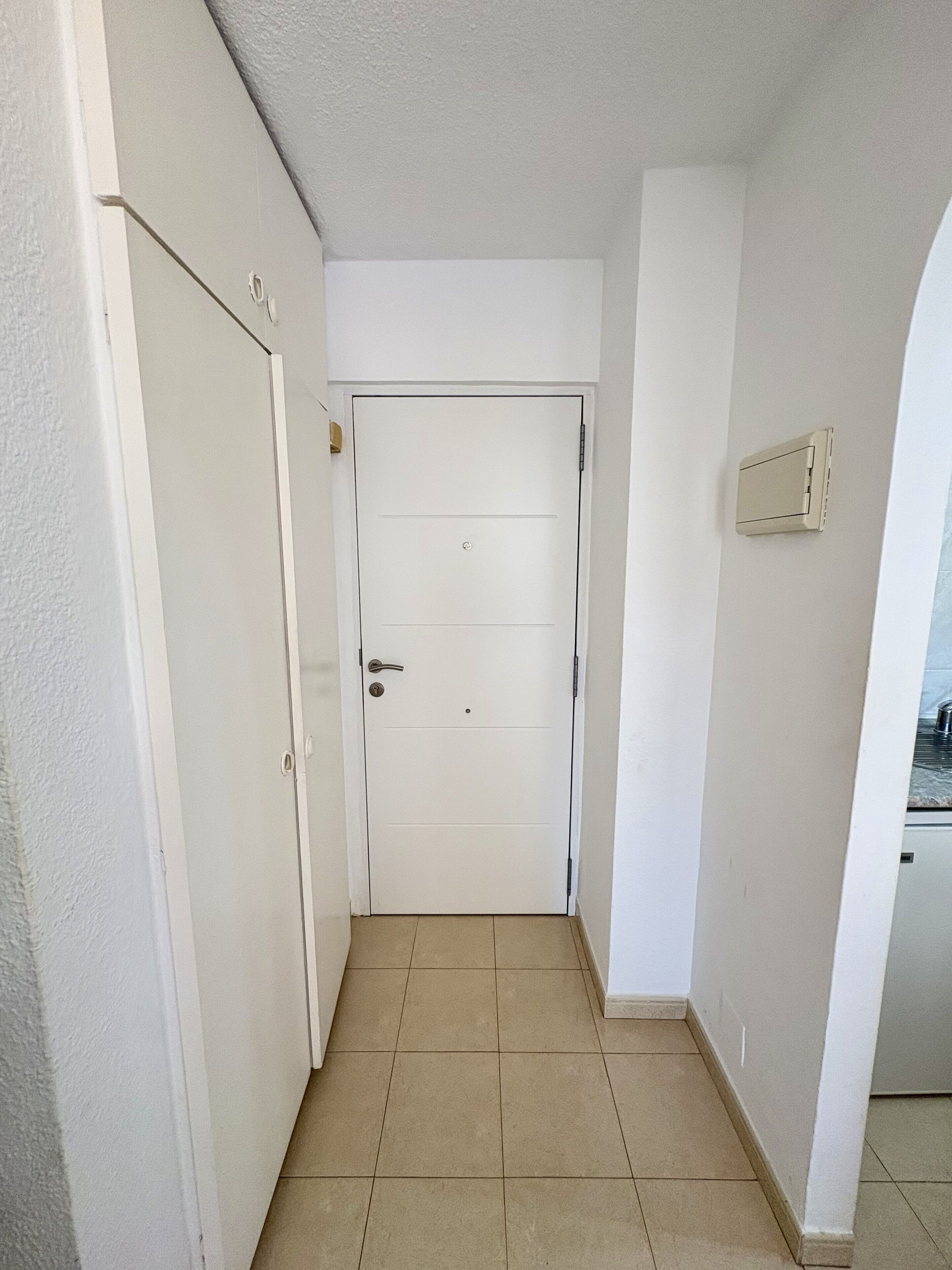 Un pasillo estrecho con una puerta blanca, un interruptor de luz en la pared y suelo de baldosas.
