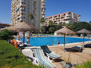 Piscina con sillas de descanso y sombrillas, rodeada de edificios altos.